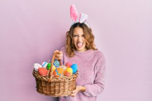 Young caucasian woman wearing cute easter bunny ears holding colored egg sticking tongue out happy with funny expression.